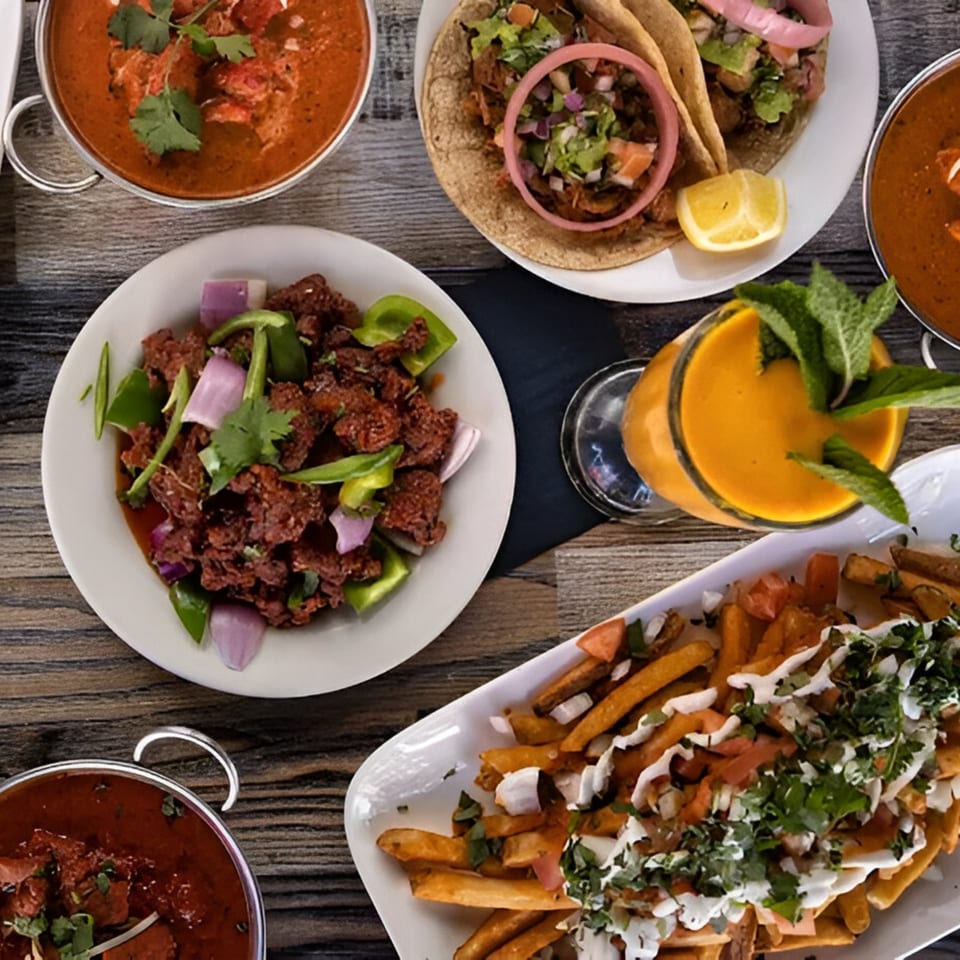 Overhead view of Indian dishes: curries, tacos, chili chicken, loaded fries, and a mango lassi.