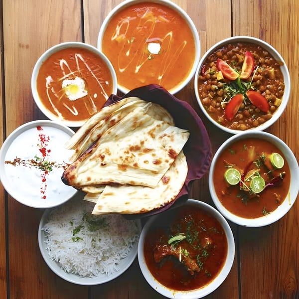 Overhead view of a variety of Indian dishes, including curries, naan, rice, and raita, on a wooden table.